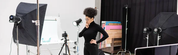 Pensativo gerenciador de conteúdo americano africano em pé perto de câmera digital e equipamentos de iluminação em estúdio de fotos espaçoso, banner — Fotografia de Stock