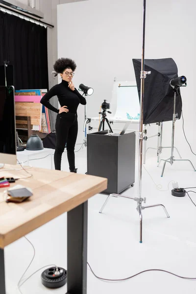Pensativo Africano americano fabricante de conteúdo de pé com a mão no quadril perto de laptop e softbox refletor no estúdio de fotos — Fotografia de Stock