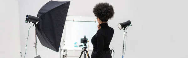 Vista posterior del administrador de contenido afroamericano en ropa negra cerca de equipos de iluminación y cámara digital en el estudio de fotografía, pancarta - foto de stock