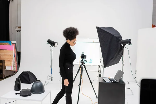 Vista lateral del productor de contenido afroamericano en ropa negra cerca de la computadora portátil y equipo de iluminación en el moderno estudio de fotografía - foto de stock