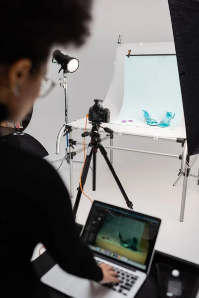 Producteur de contenu afro-américain flou travaillant sur un ordinateur portable près de l'appareil photo numérique et des chaussures à la mode sur la table de tir dans un studio photo — Photo de stock