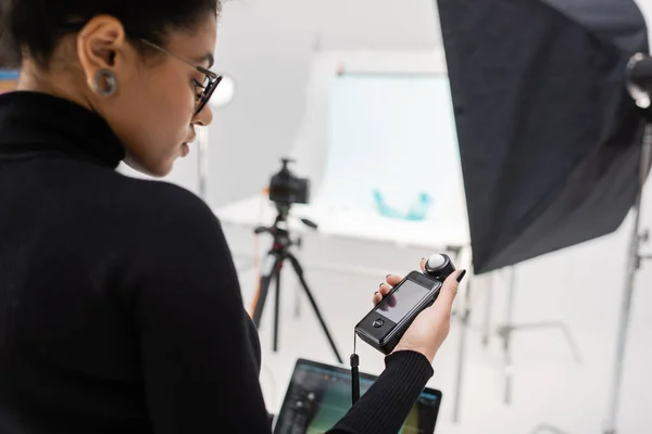 Producteur de contenu afro-américain à col roulé noir regardant le compteur d'exposition dans un studio photo flou — Photo de stock