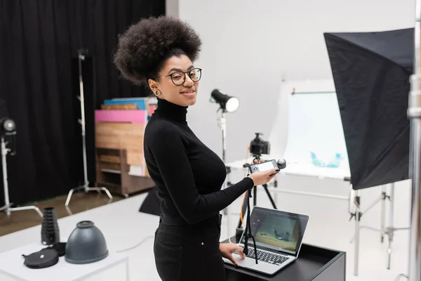 Feliz fabricante de conteúdo americano africano segurando medidor de exposição e olhando para a câmera perto do laptop no estúdio de fotos — Fotografia de Stock