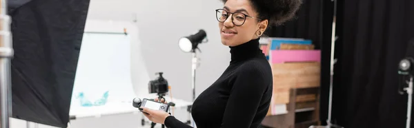 Positive african american content maker in eyeglasses holding exposure meter and smiling at camera in photo studio, banner — Stock Photo