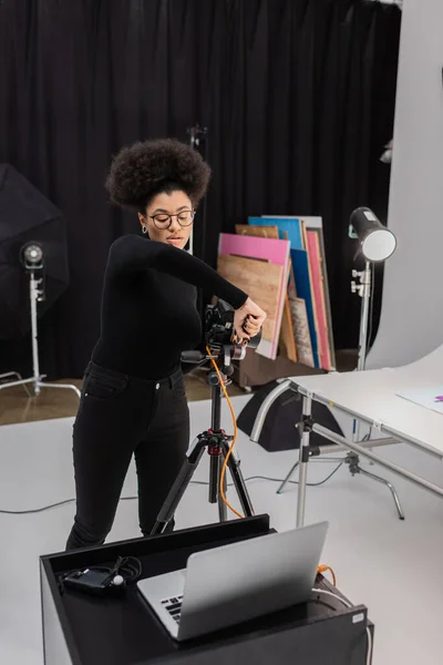 Produtor de conteúdo afro-americano em óculos montando câmera digital em tripé perto de laptop em estúdio de fotos — Fotografia de Stock