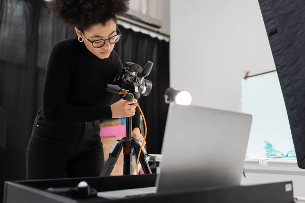 Productor de contenido afroamericano ajustando la cámara digital en el trípode cerca de la computadora portátil borrosa en el estudio de fotos - foto de stock