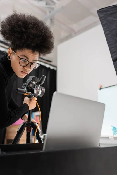 Productor de contenido afroamericano en gafas que trabajan cerca de la computadora portátil y la cámara digital en el trípode en el estudio de fotos - foto de stock