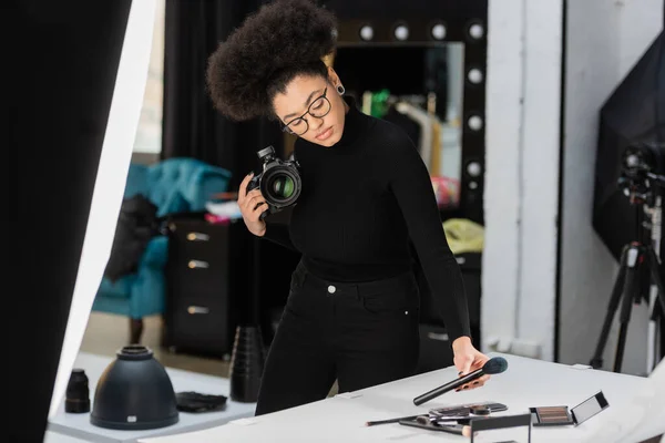 Fabricante de conteúdo americano africano com câmera digital e escova cosmética perto da mesa de tiro no estúdio de fotos — Fotografia de Stock
