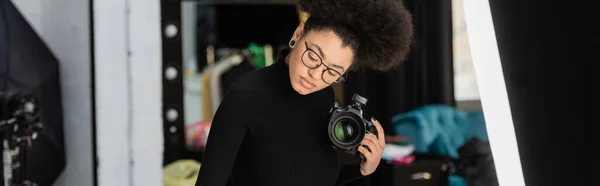African american photographer in eyeglasses holding professional digital camera in photo studio, banner — Stock Photo
