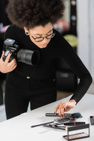 Productor de contenido afroamericano con cámara digital colocando cepillo cosmético cerca de cosméticos decorativos en la mesa de fotos en el estudio de fotografía - foto de stock