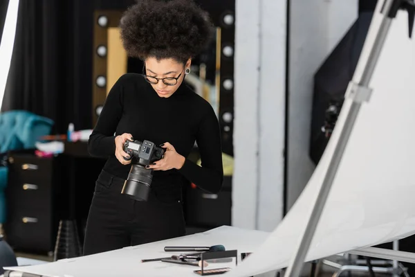 African american content manager looking at digital camera near decorative cosmetics and beauty tools in photo studio — Stock Photo