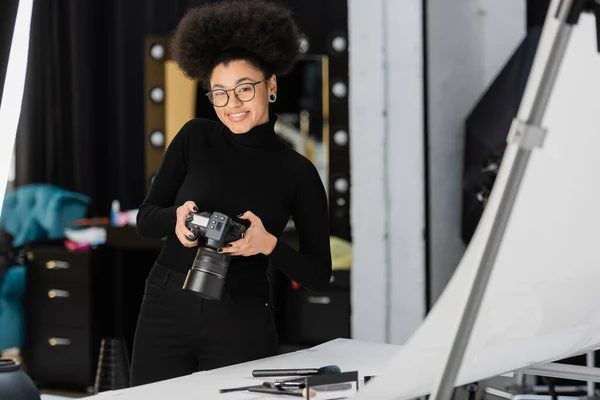 Produtor de conteúdo americano africano alegre com câmera digital sorrindo perto de cosméticos decorativos na mesa de tiro no estúdio de fotos — Fotografia de Stock