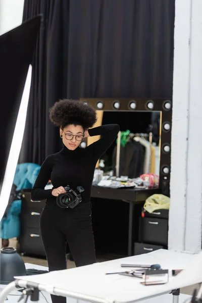 Вдумливий афроамериканський виробник контенту з цифровою камерою, посміхаючись і дивлячись на декоративну косметику на знімальному столі в фотостудії — стокове фото