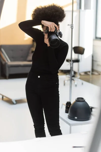 Афроамериканський виробник контенту в чорному одязі фотографується на професійній цифровій камері під час роботи в фотостудії — стокове фото
