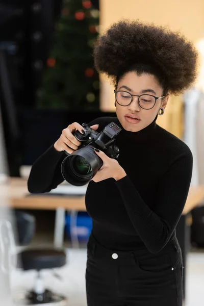 Elegante fotógrafo afroamericano en gafas y cuello alto negro trabajando con cámara digital en estudio fotográfico - foto de stock