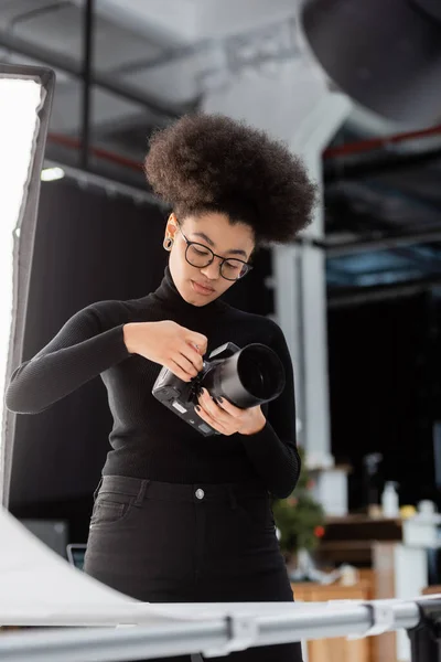 Fotógrafo afroamericano en cuello alto negro y anteojos ajustando la configuración de la cámara digital en el estudio de fotografía - foto de stock