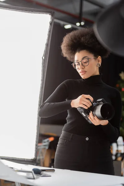 Stylish african american content maker with digital camera looking at decorative cosmetics on shooting table in photo studio — Stock Photo