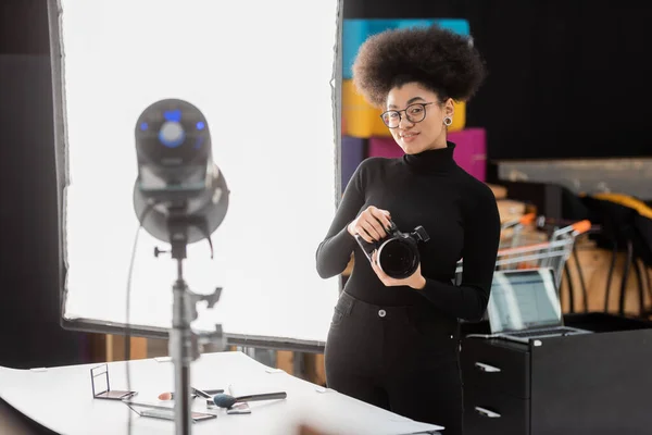 Cheerful african american content maker with digital camera smiling near blurred spotlight in photo studio — Stock Photo