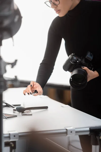 Partial view of african american content producer with digital camera and mascara near decorative cosmetics on shooting table in photo studio — Stock Photo