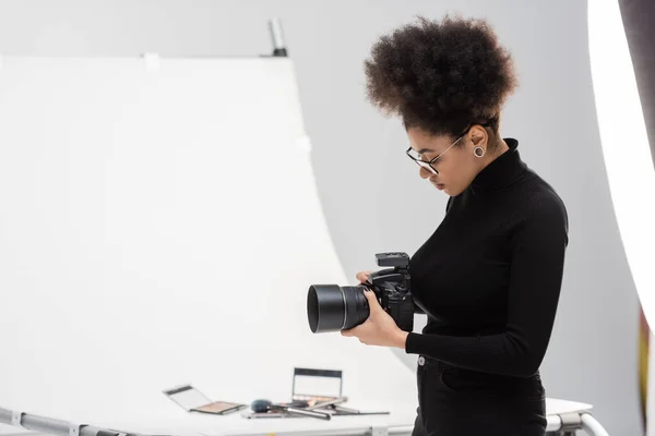 Side view of african american photographer with digital camera near shooting table with decorative cosmetics in photo studio — Stock Photo