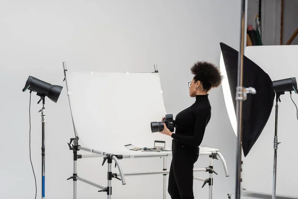 Вид збоку афроамериканського виробника контенту з цифровою камерою, що дивиться на декоративну косметику та інструменти краси в фотостудії — Stock Photo