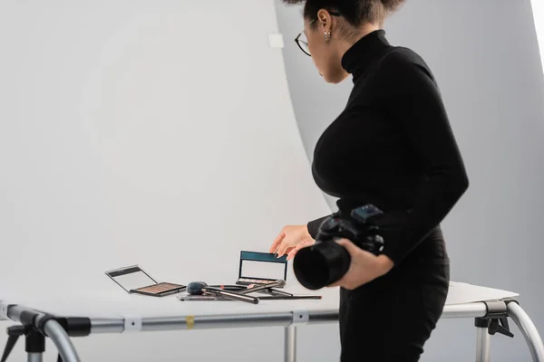 African american content maker with digital camera touching eye shadows palette near decorative cosmetics on shooting table in photo studio — Stock Photo