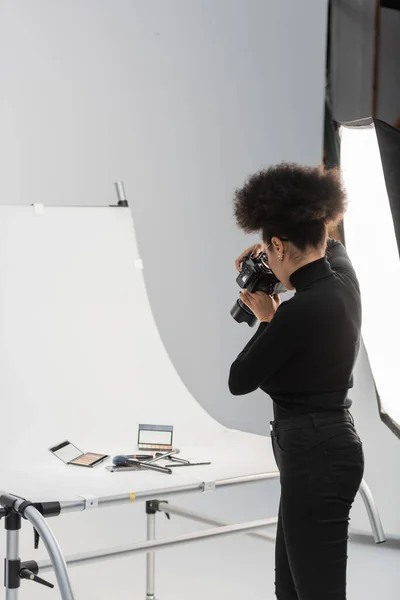 Photographe afro-américain à col roulé noir prenant des photos de cosmétiques décoratifs sur table de tir en studio photo — Photo de stock