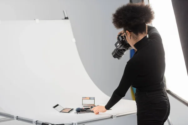 African american photographer taking photo of beauty tools and decorative cosmetics on shooting table in photo studio — Stock Photo