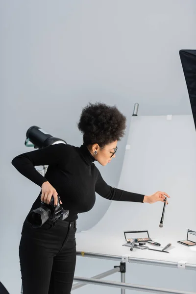 Producteur de contenu afro-américain tenant un appareil photo numérique et une brosse cosmétique près de la table de tournage dans un studio photo — Photo de stock