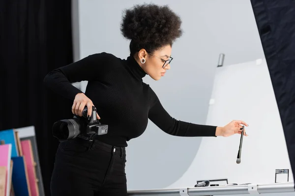 Стильний афроамериканський виробник контенту тримає косметичний пензлик біля декоративної косметики на знімальному столі в фотостудії — стокове фото