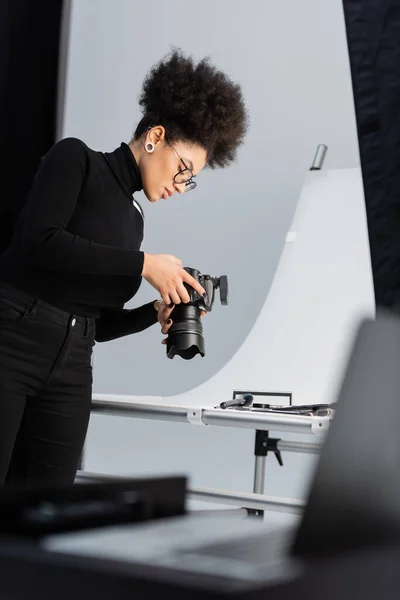 African american photographer in black turtleneck and eyeglasses looking at digital camera near decorative cosmetics in photo studio — Stock Photo