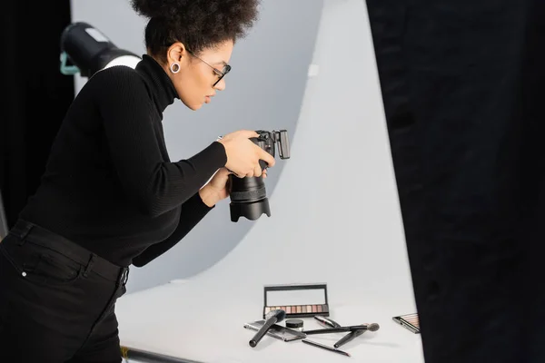 Side view of african american photographer in black turtleneck taking picture of beauty tools and cosmetics on shooting table — Stock Photo