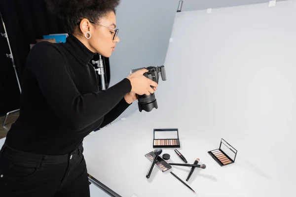 Fotógrafo afro-americano tirar foto de cosméticos decorativos e ferramentas de beleza na mesa de tiro no estúdio de fotos — Fotografia de Stock