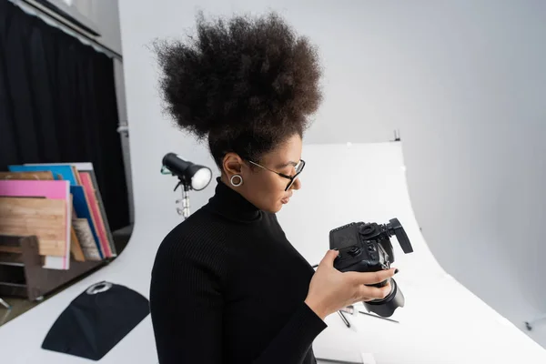 Vista lateral del fotógrafo afroamericano mirando la pantalla de la cámara digital en el estudio de fotografía - foto de stock