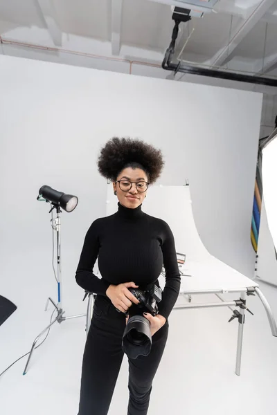 Gioioso gestore di contenuti africani americani con fotocamera digitale vicino ai riflettori e tavolo da ripresa nel moderno studio fotografico — Foto stock
