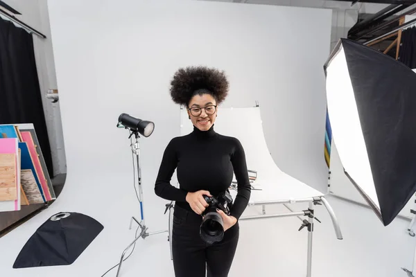 Fotografo afroamericano spensierato in dolcevita nero e occhiali da vista guardando la fotocamera vicino ai riflettori nel moderno studio fotografico — Foto stock