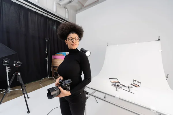 Felice creatore di contenuti africani americani con fotocamera digitale guardando lontano vicino cosmetici decorativi sul tavolo da ripresa in studio fotografico — Foto stock