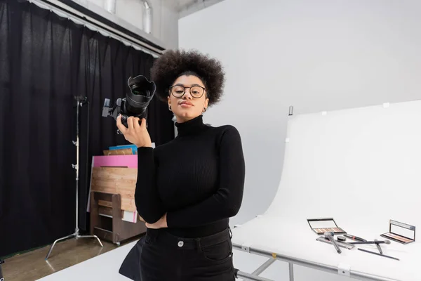 Усміхнений афроамериканський виробник контенту тримає цифрову камеру, стоячи за зйомкою столу з декоративною косметикою в фотостудії — стокове фото