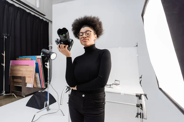 Positive african american content maker in black turtleneck and eyeglasses standing with professional digital camera in photo studio — Stock Photo