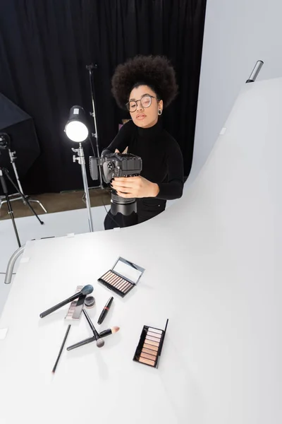 Fotógrafo afro-americano em óculos tirando fotos de ferramentas de beleza e cosméticos na mesa de tiro no estúdio de fotos — Fotografia de Stock