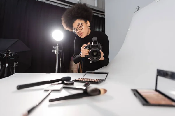 Gerente de contenido afroamericano en gafas que toman fotos de herramientas de belleza borrosas y cosméticos decorativos en el estudio de fotografía - foto de stock