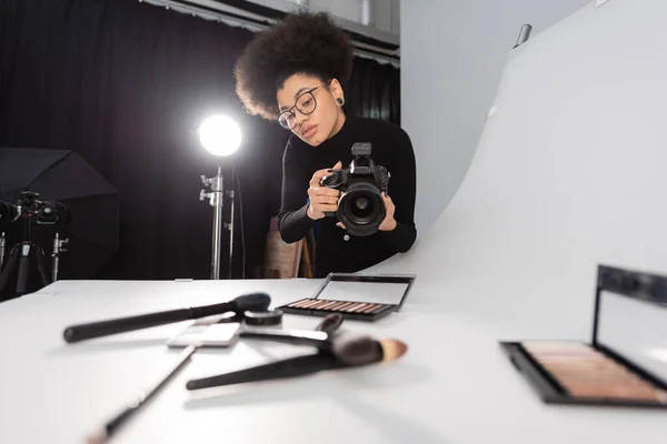 Gestionnaire de contenu afro-américain en lunettes prenant des photos d'outils de beauté flous et de cosmétiques décoratifs dans un studio photo — Photo de stock