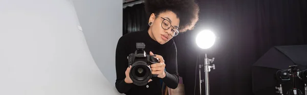 Producteur de contenu afro-américain avec appareil photo numérique travaillant près de projecteur dans un studio photo, bannière — Photo de stock