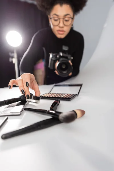 Blurred african american content producer holding cosmetic cream near eye shadows and brushes on shooting table in photo studio — Stock Photo