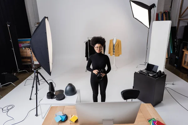 Lächelnder afrikanisch-amerikanischer Fotograf in der Nähe von Scheinwerfern und einer neuen Kollektion trendiger Kleidung im modernen Fotostudio — Stockfoto