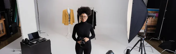 African american content maker with digital camera near collection of stylish clothes and laptop in photo studio, banner — Stock Photo