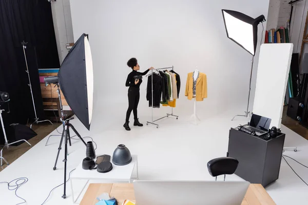 Longitud completa del productor de contenido afroamericano que trabaja con la nueva colección de ropa de moda cerca de focos en el estudio de fotos - foto de stock