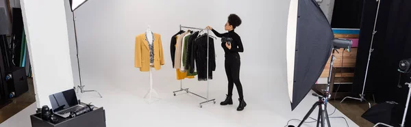 Comprimento total do fabricante de conteúdo americano africano perto de nova coleção de roupas elegantes e holofotes no estúdio de fotos, banner — Fotografia de Stock