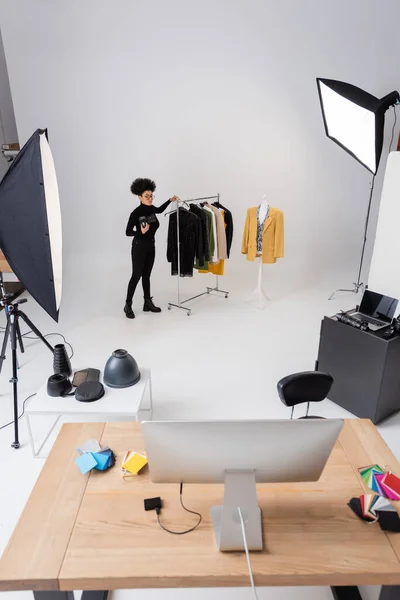 Full length of african american content manager near rail rack with trendy clothing in photo studio with computers and lighting equipment — Stock Photo