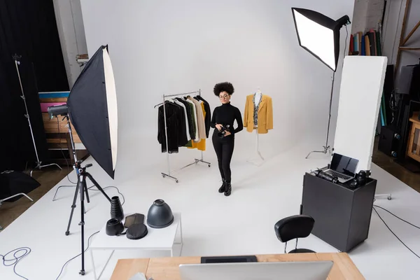 Comprimento total do produtor de conteúdo americano africano com câmera digital perto de roupas da moda em estúdio de fotos espaçoso com equipamento de iluminação — Fotografia de Stock